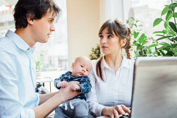 Happy two parents looking at each other, father holding newborn baby boy, mother working with tablet computer.