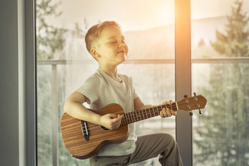 Young boy play on guitar at home at sunny day. Boy play on ukule