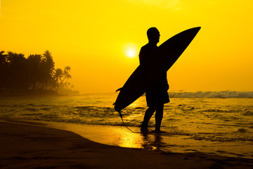 The male figure with a board for surf on a background of ocean waves. The setting sun paints the sky in bright orange yellow gold color