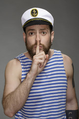 No words. Waist up shot of an attractive bearded man wearing sailor outfit making shushing gesture with his finger to his lips on grey background.