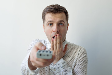 Naklejka premium A surprised young man switches channels by clicking on the buttons. Shock from the information on TV.
