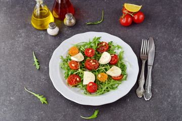 Traditional Italian salad with cherry tomato, ruccola, mozzarella, olive oil wine vinegar on a white plate on a grey abstract background.