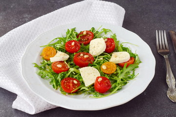 Traditional Italian salad with cherry tomato, ruccola, mozzarella, olive oil wine vinegar on a white plate on a grey abstract background.