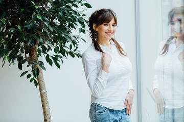 Young brunette business woman in white shirts standing near big window with reflection. Female with Scythes looking at the camera.