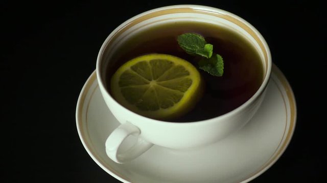 Mint leaves fall into a cup of black tea on a black table. Slow motion