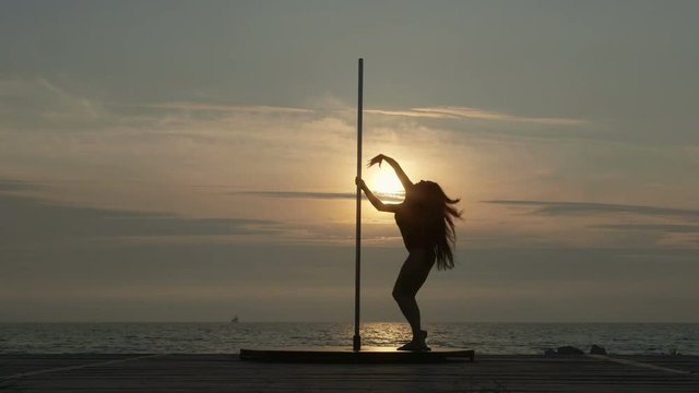 Pole dance fitness exercise on the beach. Silhouette of young woman pole dancer during outdoor workout against the setting sun.
