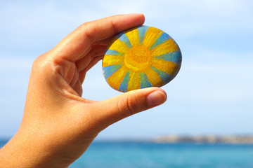 Woman hand hold pebble with painted bright sun against blue sky and sea