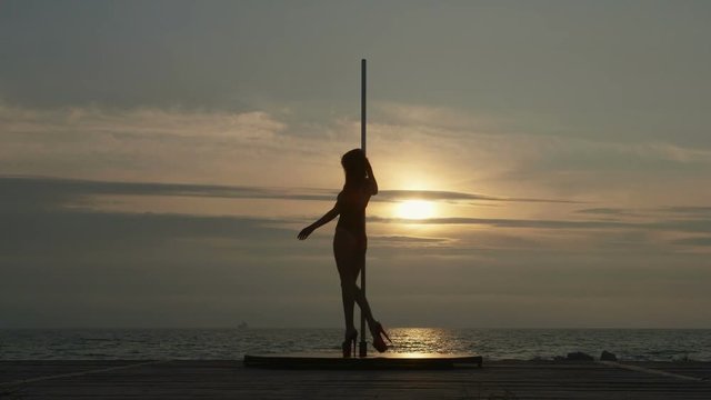 Pole dance fitness exercise on the beach at sunset. Silhouette of young woman pole dancer in high heels against the setting sun