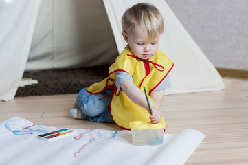 The kid in a yellow waistcoat draws a brush on a white paper with watercolors