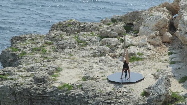 Outdoor pole dance fitness exercise above the sea. Fit girl pole dancer in bodysuit dancing on the edge of a cliff