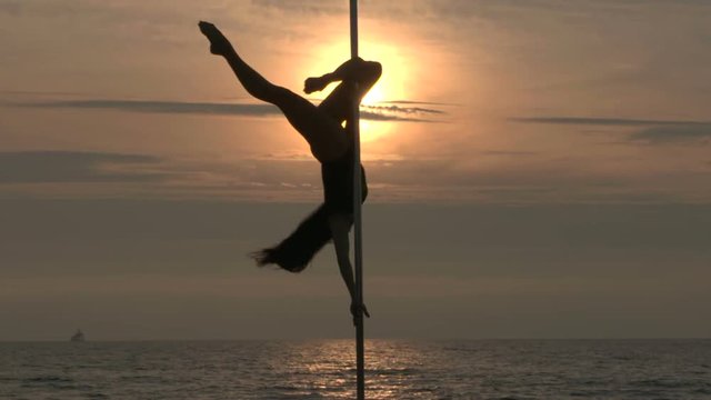 Pole dance fitness exercise on the beach at sunset. Silhouette of young woman pole dancer spinning in the air against the sun