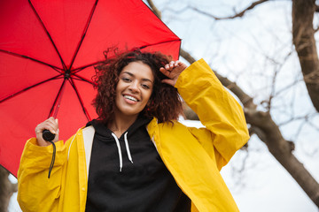 Happy african curly young lady wearing yellow coat holding umbrella