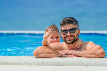 Obraz premium Seven years old cute boy smiling without tooth. He and his bearded father are in sunglasses, show their tongues, they leaned on pool railing giving a grimace of happiness. Sea view and beach umbrellas