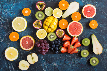 Variety of colourful fruits, oranges, lemons, kiwis, grapefruits, strawberries, grapes, blueberries, pears, passion fruit, mango on the black slate on the dark blue background, top view