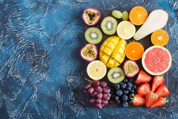 Fototapeta na wymiar Top view of different raw fruits platter: oranges, lemons, kiwis, grapefruits, strawberries, grapes, blueberries, pears, passion fruit, mango on the black slate on the dark blue background