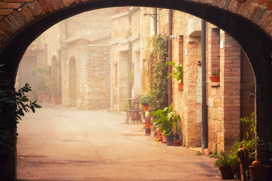 Fototapeta Narrow street of medieval San Quirico d'Orcia city with arch, green plants and cobblestone, travel foggy morning Italy background