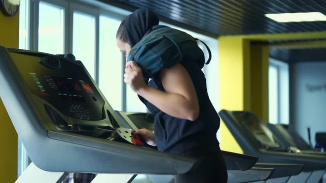 A young athlete is engaged on a treadmill in a fitness club, changing the intensity of the run. A woman is running with a bag on her shoulders to increase the load on her legs.