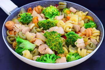 Stir fried chicken fillet with vegetables and pasta in a frying pan on a black abstract background.