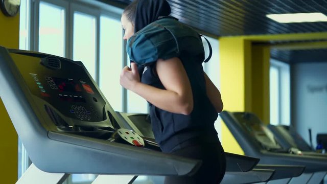 Pretty sporty woman is engaged on a treadmill in a fitness club, changing the intensity of the run. A woman is running with a bag on her shoulders to increase the load on her legs.