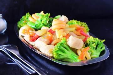 Stir fried chicken fillet with vegetables and pasta on a metal tray on a black abstract background.