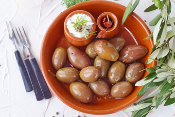 Olives in a traditional brown ceramic bowl, with tzatziki sauce, lemon, garlic, olive branch and olive oil on a white abstract background.