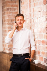 Handsome businessman in white shirt is speaking on the phone near the brick wall. There is a cup of coffee nearby