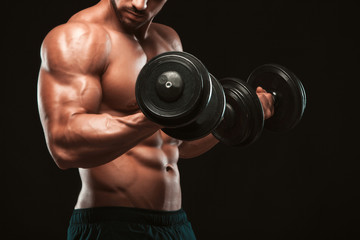 Handsome power athletic man in training pumping up muscles with dumbbells in a gym. Fitness muscular body isolated on dark background.