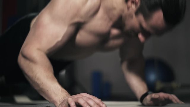 Close up of a young bearded man with long hair and bare torso doing push ups in a gym. Locked down real time close up shot.