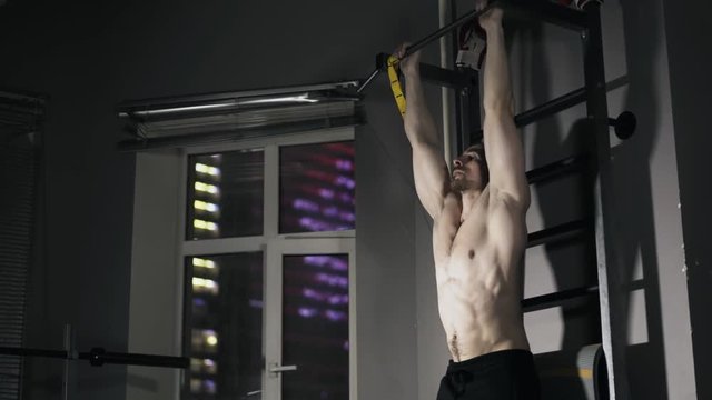 Tilt down of a young bearded man with a bare torso doing leg lifting on a horizontal bar in a gym. Locked down real time medium shot