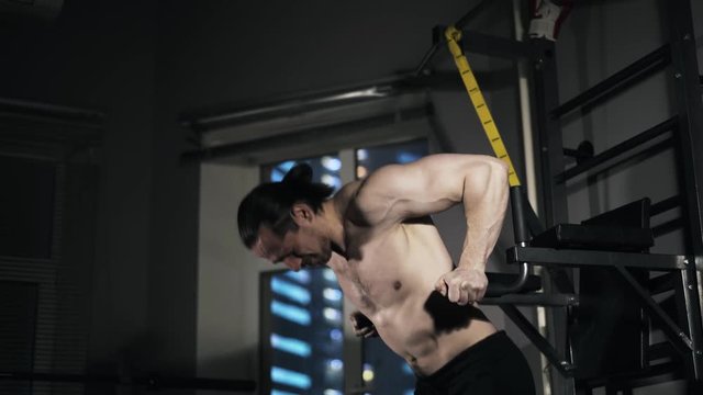 Bearded young man with a bare torso is doing push ups on parallel bars in a gym and walking away. Locked down real time medium shot.