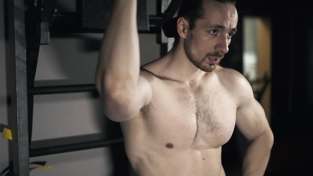 Front view of a bearded young bodybuilder with a naked torso breathing heavily after an intensive workout in a gym. Real time tilt up medium shot