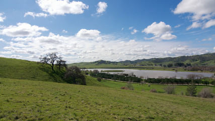 Fototapeta na wymiar Panoramic view of the Lagoon Valley Park in Vacaville, California, USA, featuring the chaparral in the winter with green grass, and the lake, with interesting clouds in the sky