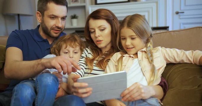 Mother, father, son and daughter using tablet, watching movie, playing games, tapping and scrolling touchscreen. Family spending time together at home. Happy family