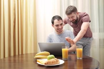 Happy gay couple having breakfast in kitchen and using laptop
