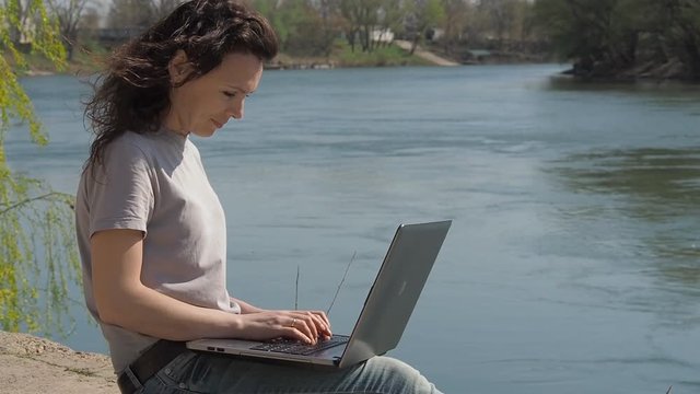 Young woman at the river with a laptop. Girl sits with a laptop on the river bank. Slow motion.