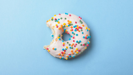 bitten donut with white glaze and colorful sprinkles isolated on blue background. top view