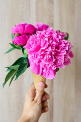 white caucasian women's hand keeping ice cream's imitation waffle cone with composition peony flowers inside house on a wooden backdrop