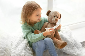 Cute little girl with teddy bear sitting on window sill at home