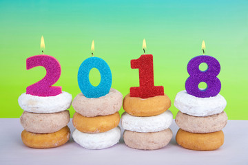 Donuts stacked with candles celebrating Happy New Year, 2018 burning bright. Blue green yellow textured background. Cake, cinnamon and powdered doughnuts in piles.
