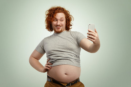 Obese young male with curly ginger hair and beard holding mobile phone, posing for selfie, looking at camera with flirty smile while his fat belly hanging out of grey shrunk t-shirt and jeans pants