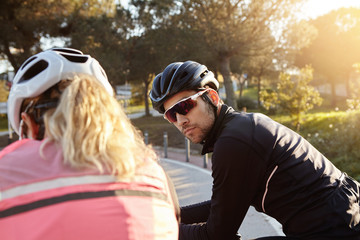 People, health, sports, fitness and active lifestyle. Attractive young cyclist in sunglasses and helmet having conversation outdoors with his blonde girlfriend, who is standing with her back to camera