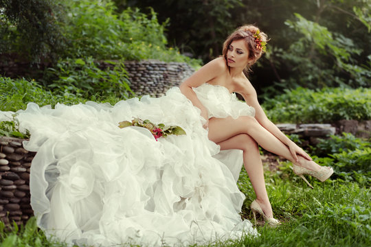 Sexy bride woman with long legs in lush wedding dress sits on nature.