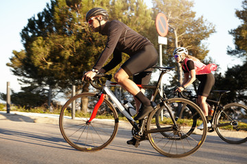 Sideways outdoor shot of two young people wearing protective gear and cycling clothing riding racing bicycles uphill on city road, enjoying warm sunny morning. Sports, fitness and healthy lifestyle