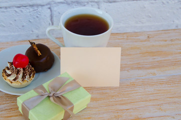 Cup of tea with cute sweet cakes , gift box and  greeting card with space for text on wooden background. Mothers day breakfast concept.