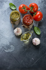 Glass jars with red and green pesto and cooking ingredients, dark grey stone background, copyspace