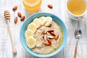 Bowl of oatmeal porridge with bananas, honey, almonds and sunflower seeds and cup of green tea on white table, closeup view, top view. Concept of dieting, healthy breakfast, healthy eating