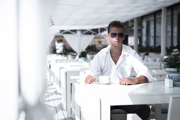 Young serious business style male is drinking coffee in seaside restaurant. Sitting alone wearing stylish sunglasses and white shirt Caucasian guy is looking forward at the camera.