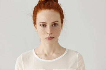 Pretty student girl with ginger hair in knot relaxing at home after college. Headshot of tender charming young woman with freckles wearing white blouse posing at studio wall, having calm expression - obrazy, fototapety, plakaty