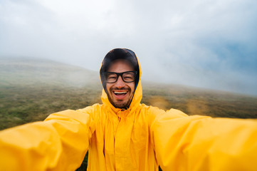 Selfie portrait of a smiling and laughing traveller man in yellow raincoat and glasses in the clouds mountains in stromy weather with rain