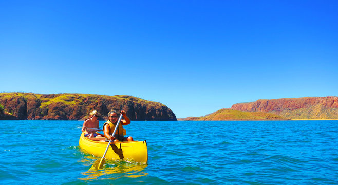 Two happy smile girls / women enjoying kayaking on the large freshwater Lake Argyle in Western Australia on a beautiful sunny day with clear blue skies and red rock mountains in the background